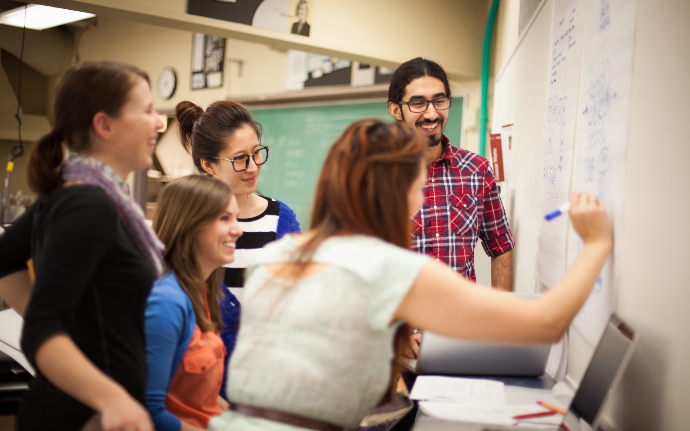 a group of five smiling students in casual clothing facing a whiteboard with one student drawing on the board with a blue marker and a laptop in the foreground