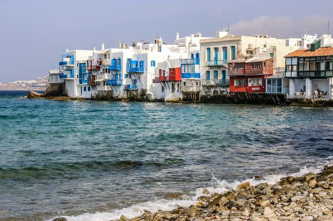 Colourful, tightly packed buildings on a shoreline in Greece, with a pebbly beach in the foreground.